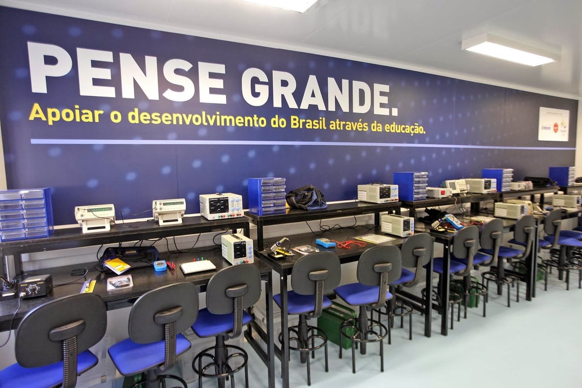 Laboratório de informática com placa azul escrito PENSE GRANDE e fileiras de computadores e cadeiras azuis